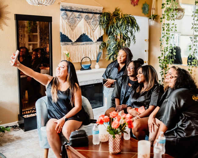 A group of six women in matching black robes pose together for a selfie in a cozy living room adorned with plants and decorative accents.