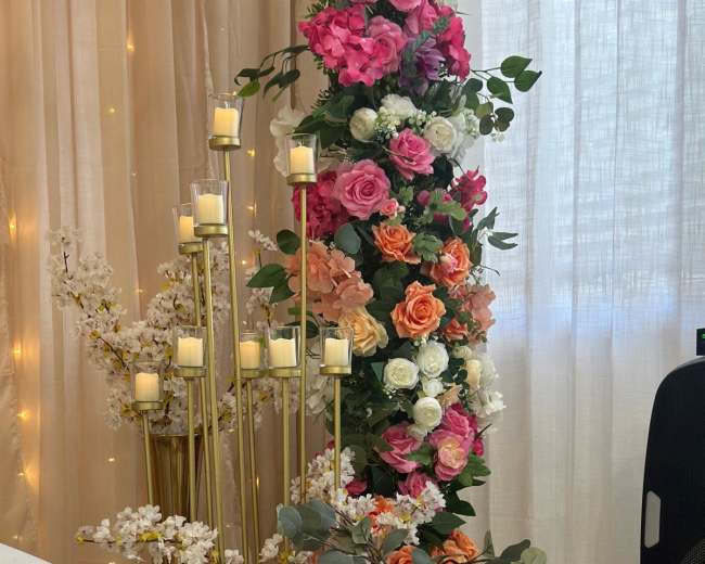 The image features a decorative arrangement of various artificial flowers in vibrant colors, alongside tall candle holders and a woven basket filled with flowers, set against a backdrop of soft drapery.