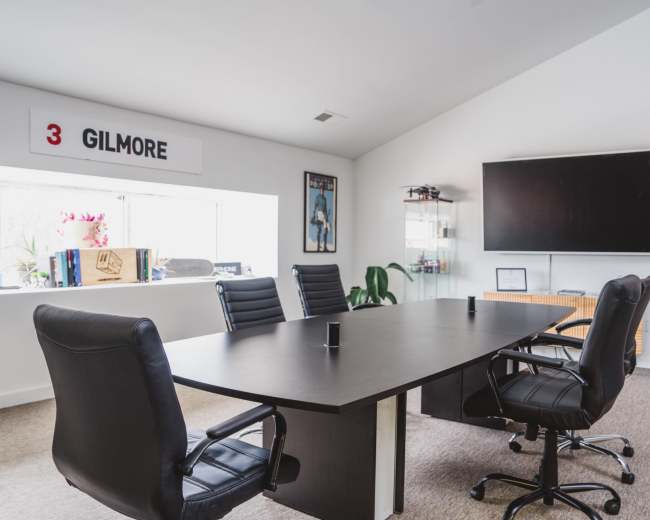 A modern conference room features a large table surrounded by black office chairs, with a window displaying the name "GILMORE" and a wall-mounted television.