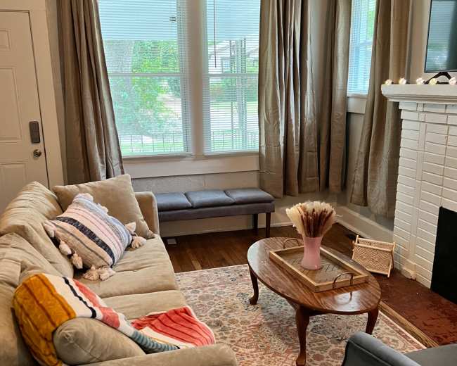 A living room with a beige sofa, a blue armchair, a wooden coffee table, and a white brick fireplace, all complemented by large windows draped with beige curtains.