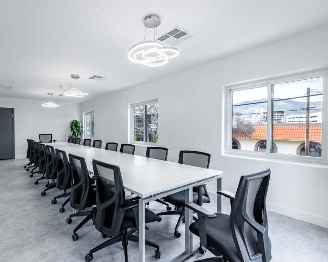 A modern conference room features a long white table surrounded by black office chairs, with large windows allowing natural light and two circular light fixtures overhead.