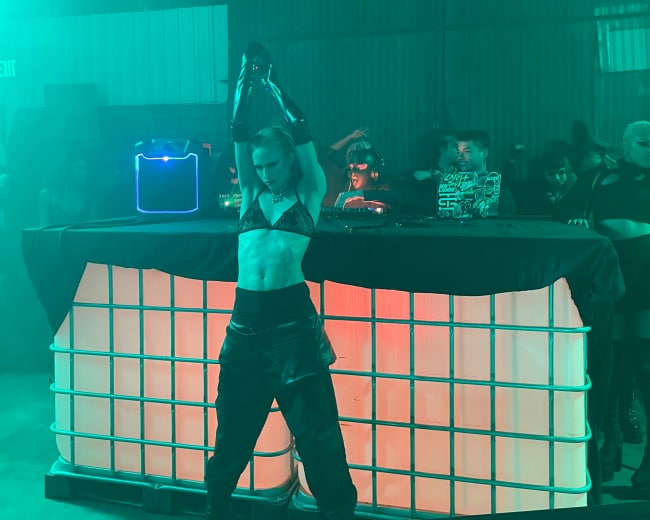 A performer in a black outfit dances at a nightclub while a DJ plays music behind a illuminated table.