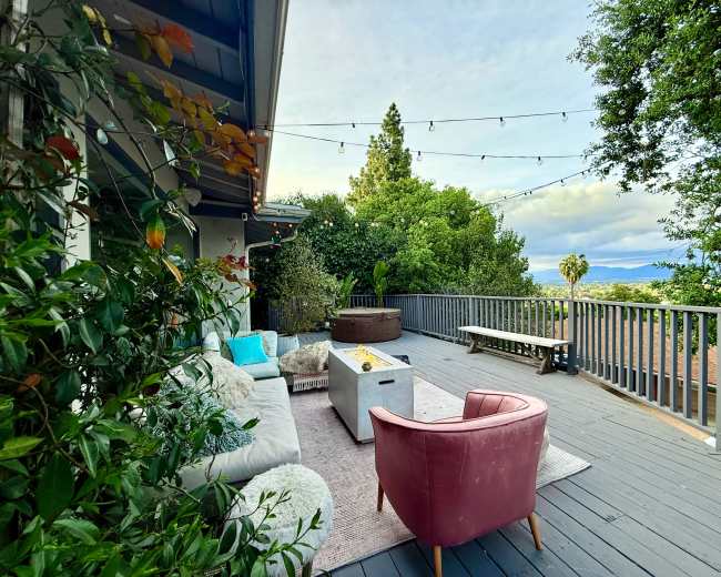 A wooden deck with a cozy seating area, potted plants, and string lights offers a view of trees and distant hills.