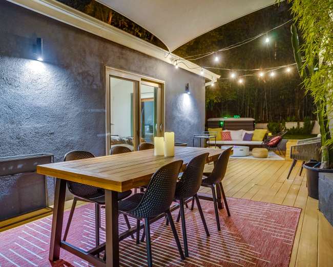 The image shows a modern outdoor dining area featuring a wooden table surrounded by black chairs, with soft lighting and a cozy atmosphere.