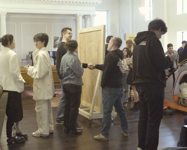 A group of people socialize and interact in a spacious room with light-colored walls and wooden floors.