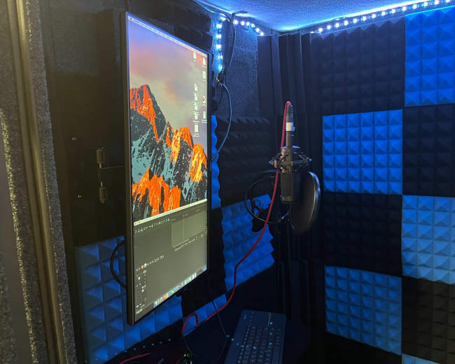 A soundproof recording booth featuring a monitor displaying editing software, a microphone mounted on a stand, and acoustic foam panels in blue and black.