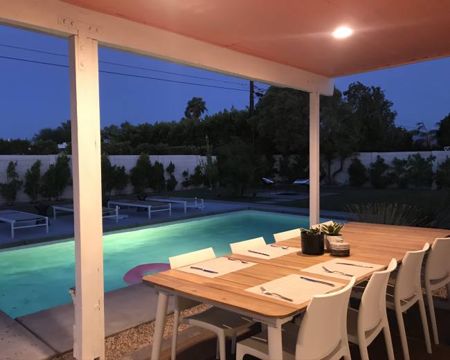 A wooden dining table with white chairs is set up under a covered patio, overlooking a swimming pool illuminated in the evening light.