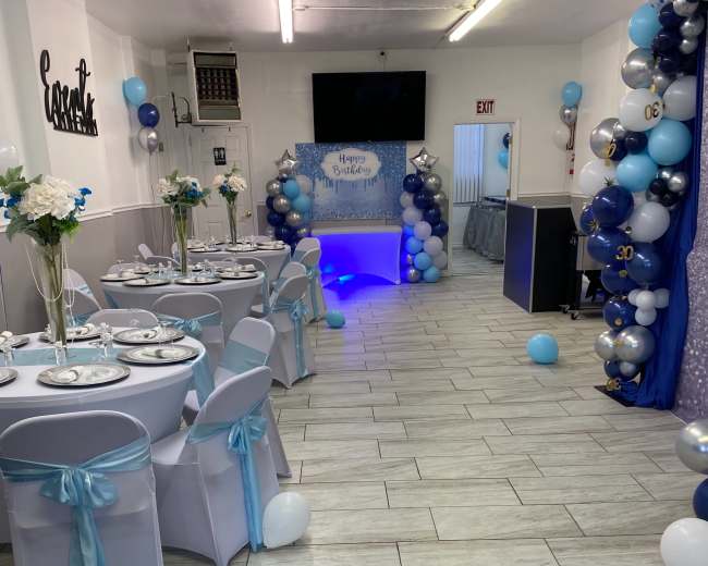 A decorated party space featuring tables with white tablecloths, blue and silver balloons, and a themed backdrop for a celebration.