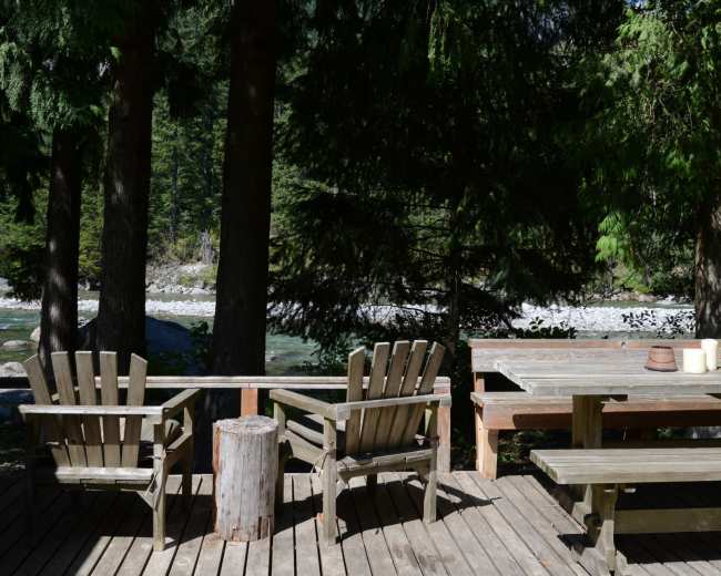 A wooden deck with two adirondack chairs and a table overlooks a river surrounded by trees.