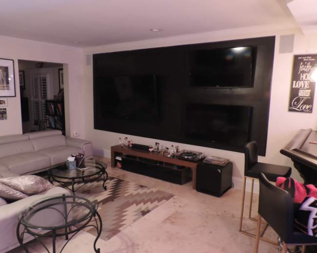 The living room features a light-colored sectional sofa, a glass coffee table, and a dark wall with three mounted flat-screen televisions.