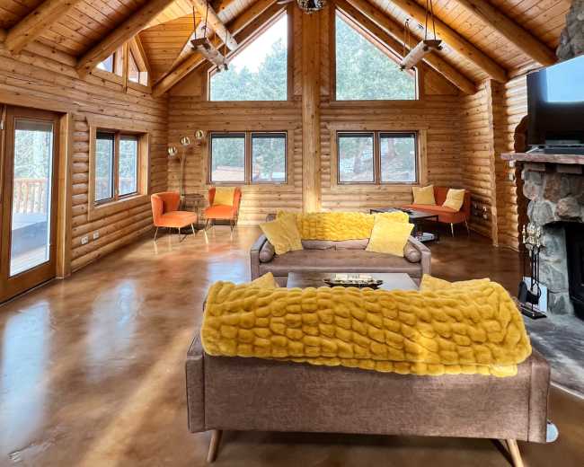 A spacious log cabin living room features a high wooden ceiling, large windows, and modern furniture arranged around a central sofa.