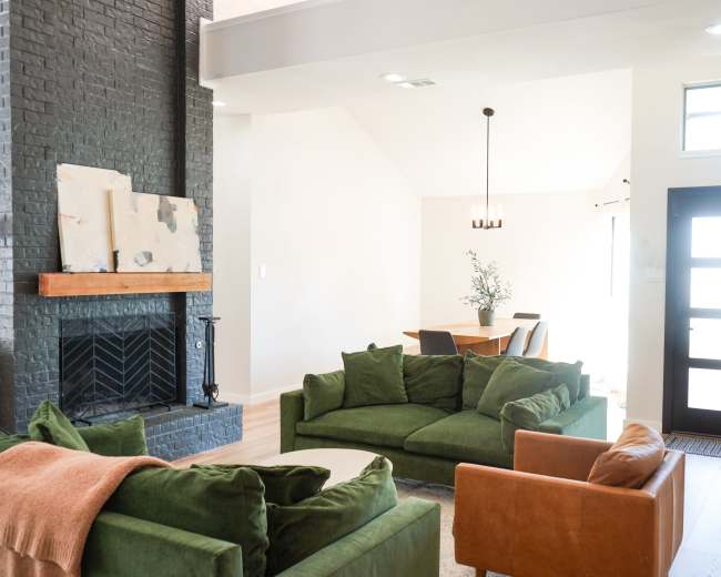 The image shows a bright living room with a dark brick fireplace, two green sofas, an orange armchair, and a dining area visible in the background.