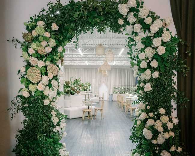 An arched entrance adorned with white hydrangeas and green leaves leads into a decorated event space with tables and floral arrangements.