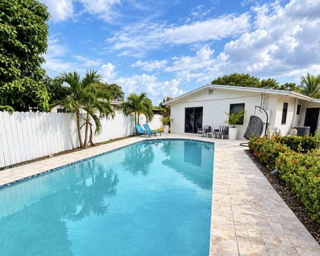 The image shows a clear, blue swimming pool surrounded by a tiled deck, tropical plants, and a white fence, with a house in the background.