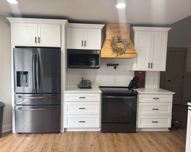 The image shows a modern kitchen featuring white cabinets, a black refrigerator, an oven, and a wooden range hood.