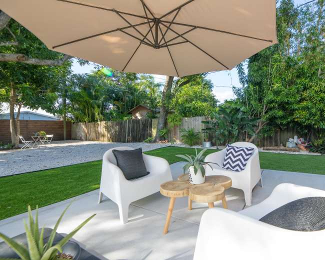 A shaded seating area features two white chairs and a wooden table on a patio surrounded by greenery and gravel in the backyard.