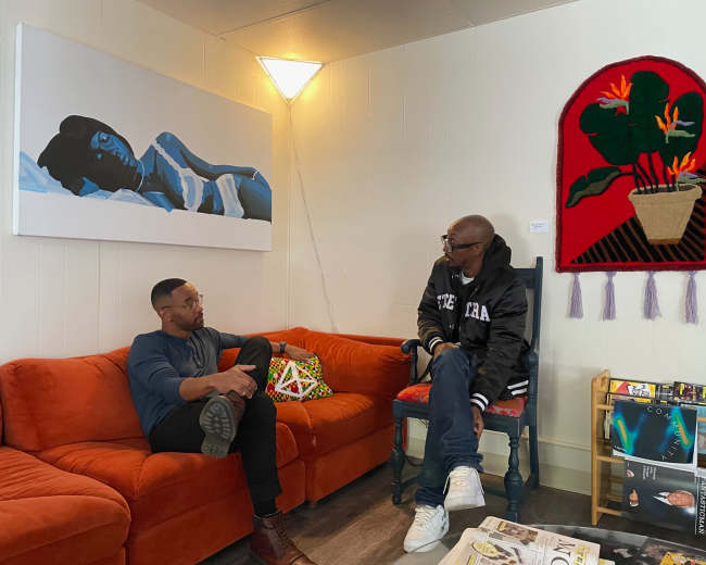 Two men are seated in a cozy room, one on a bright orange couch and the other on a chair, discussing while surrounded by artwork and decor.