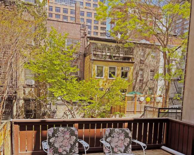 A wooden balcony with floral-patterned chairs and a table, overlooking buildings and trees in an urban setting.