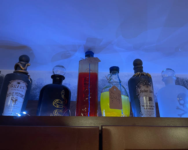 The image shows a row of decorative glass bottles, each with unique labels and colored contents, illuminated under blue lighting.