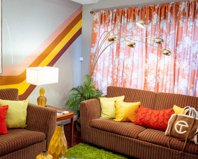 The living room features two striped sofas adorned with colorful cushions, a floor lamp, and vibrant floral curtains.