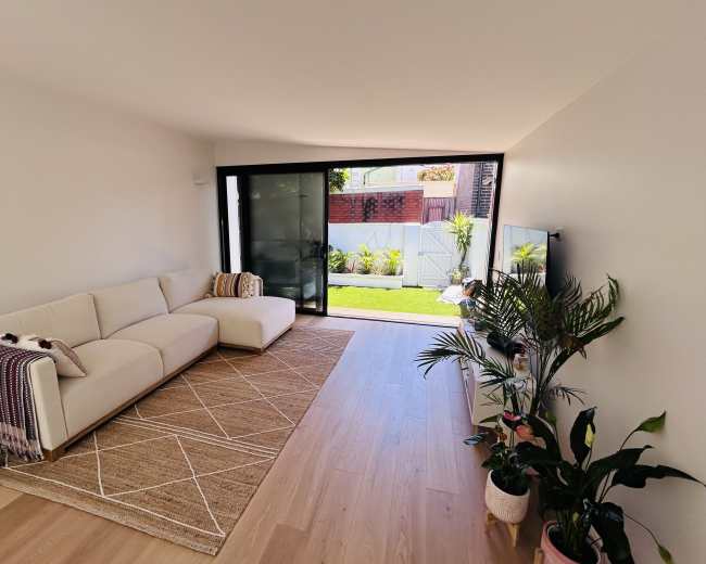 The image shows a bright living room with a white couch, a large window opening to a garden, and indoor plants.