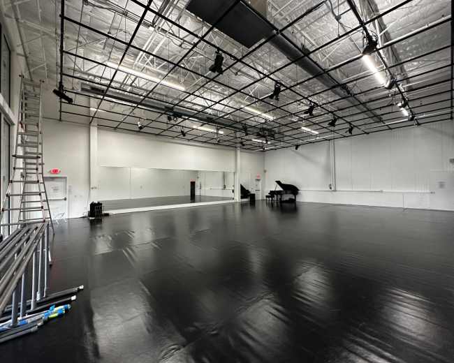 The image shows a large, empty dance studio with a glossy black floor, a mirrored wall, and a grand piano in the corner, illuminated by overhead lights.