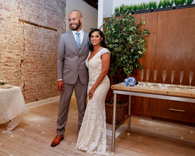 A man in a gray suit and a woman in a white lace dress stand together in a decorated venue with exposed brick walls and a table set for a celebration.