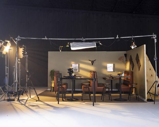 A film set is arranged with a dining area featuring two tables and chairs, surrounded by lighting equipment and cameras.