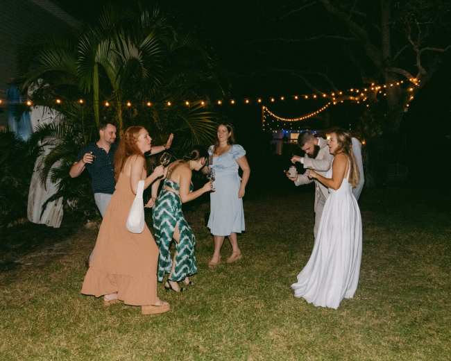 A group of people is dancing in a grassy backyard at night, with string lights illuminating the scene.