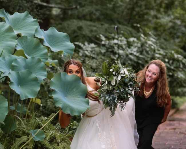 A bride holding a bouquet playfully peeks from behind a large lotus plant while a woman stands smiling nearby.