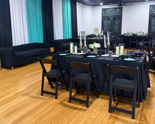 A large dining area features a black table set with candles and decorations, surrounded by black folding chairs, with teal and black drapery along the walls.