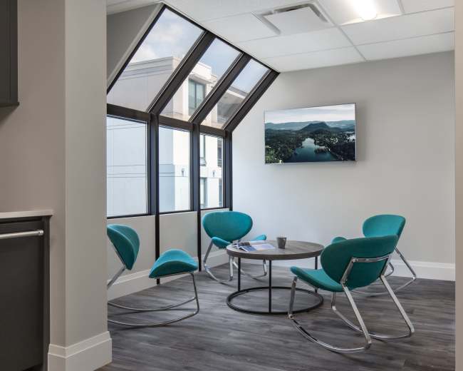 A modern waiting area features four teal chairs arranged around a circular table, with a large window providing a view of the outdoors and a television mounted on the wall.