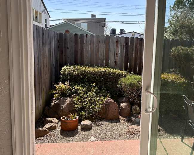 The image shows a view through a glass door into a small backyard garden with a landscaped area featuring rocks, shrubs, and a couple of chairs.
