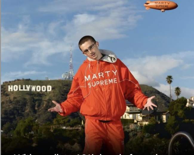 A man in an orange jumpsuit poses in front of the Hollywood sign while promoting a film related to cultural marketing.