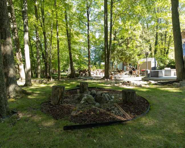 The image shows a circular fire pit area with wooden stumps arranged around it, surrounded by trees and a residential building in the background.