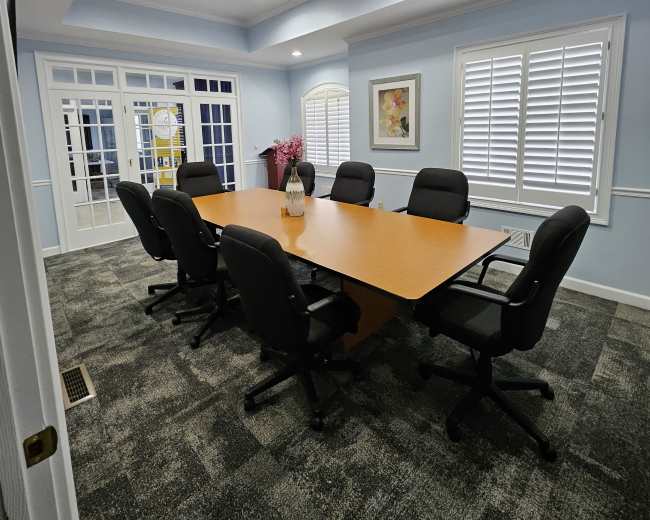 The image shows a conference room with a large wooden table surrounded by black office chairs, illuminated by recessed lighting and featuring window shutters and artwork on the walls.