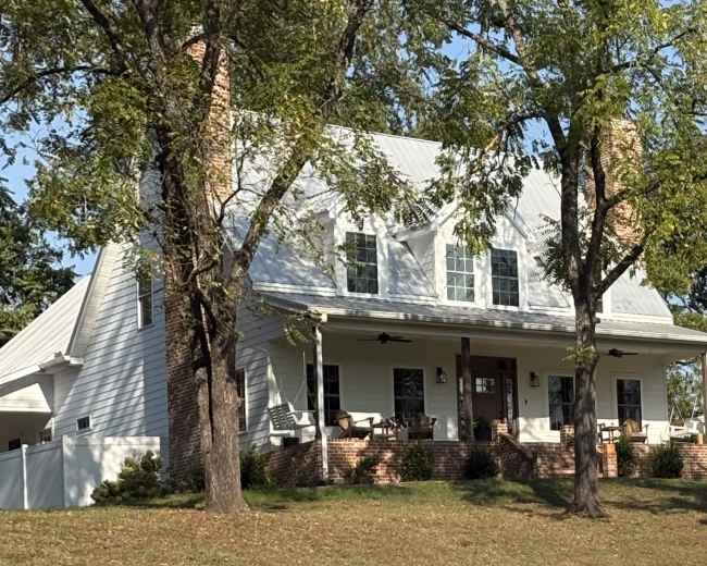 A two-story white house with a metal roof and brick foundation is surrounded by trees on a grassy slope.