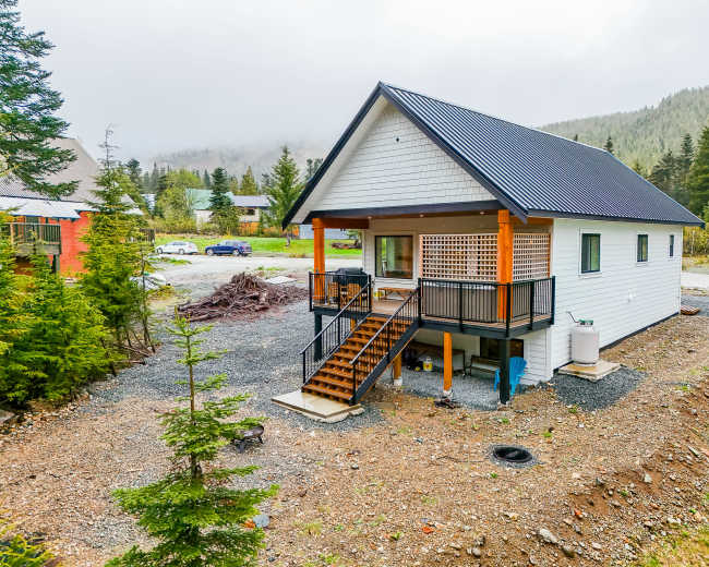 A two-story white house with a black roof is situated on a gravel lot surrounded by trees and a foggy landscape.