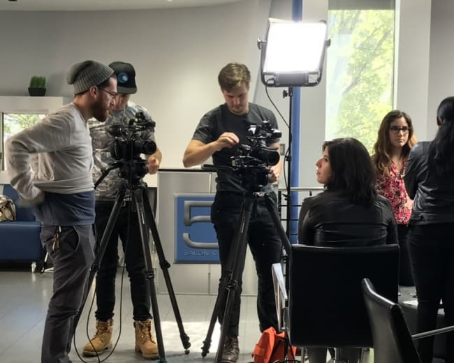 A film crew sets up equipment while a woman sits in a chair, preparing for a shoot in a modern salon.