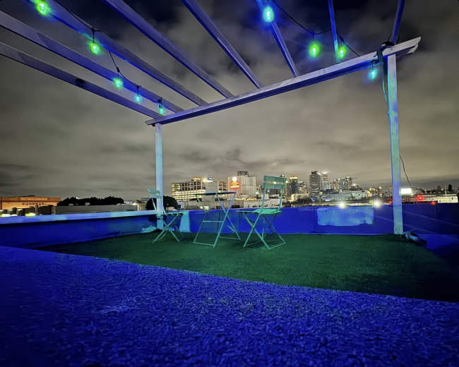 The image shows a rooftop space with green chairs under a lighted pergola, overlooking a city skyline at night.
