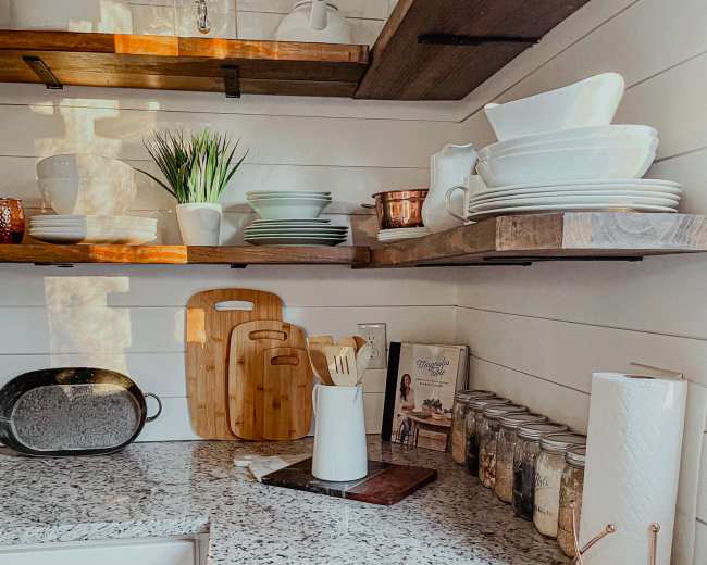 The kitchen features a granite countertop with wooden shelves above, displaying various dishware, utensils, and jars, along with a cutting board and a paper towel holder.