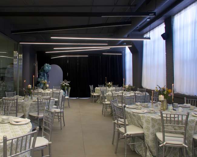 The image shows a well-arranged event space featuring round tables with floral tablecloths, silver chiavari chairs, and elegant centerpieces, illuminated by modern lighting fixtures.