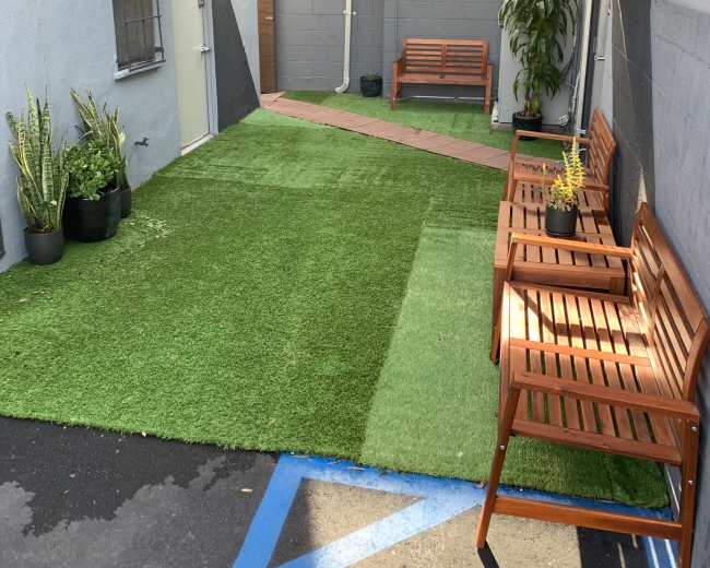 The image shows a narrow courtyard with artificial grass and wooden benches on either side, bordered by gray walls.