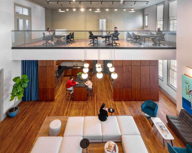 The image shows a modern office space with two levels, where people are working at desks on the upper floor and others are seated at a table on the lower floor.