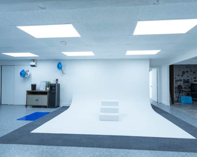 The image shows a bright, minimalist studio space with a smooth white backdrop, three white steps in the center, and a few items like a refrigerator and a desk visible in the background.