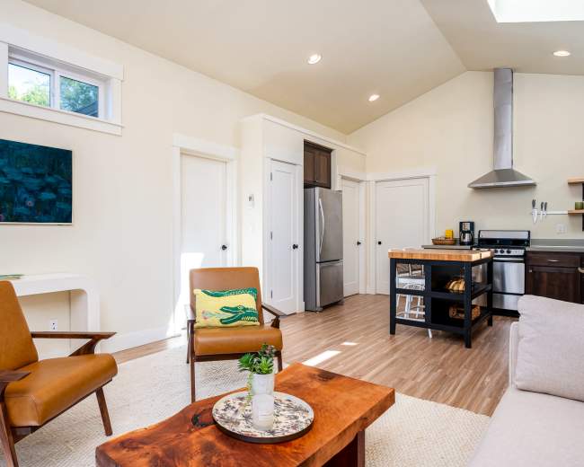 The image shows a bright, modern living space featuring a seating area with two chairs and a coffee table, and an open kitchen with stainless steel appliances.