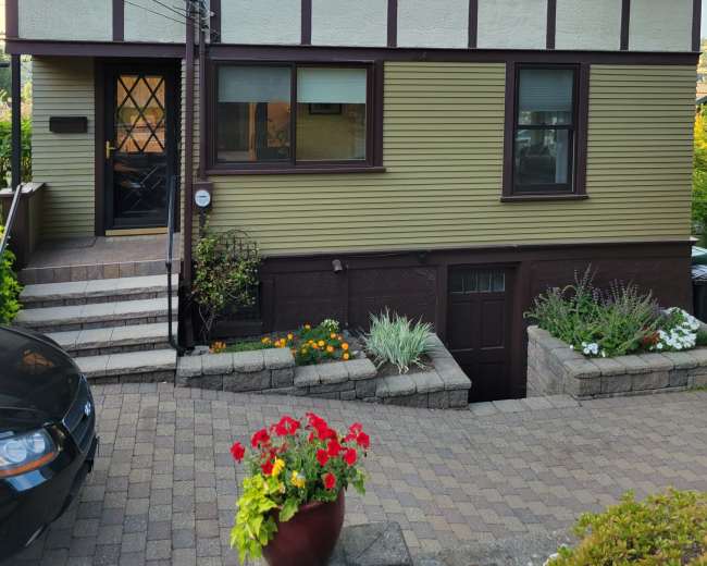 The image shows a two-story house with a combination of yellow and brown exterior, featuring a front entrance with steps and landscaped flower beds.