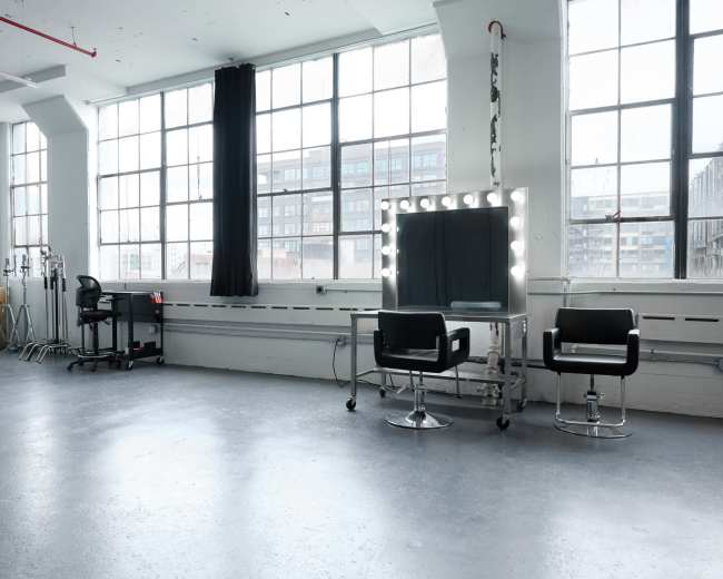 A spacious, well-lit room features large windows, a mirrored vanity with lights, and two black chairs.