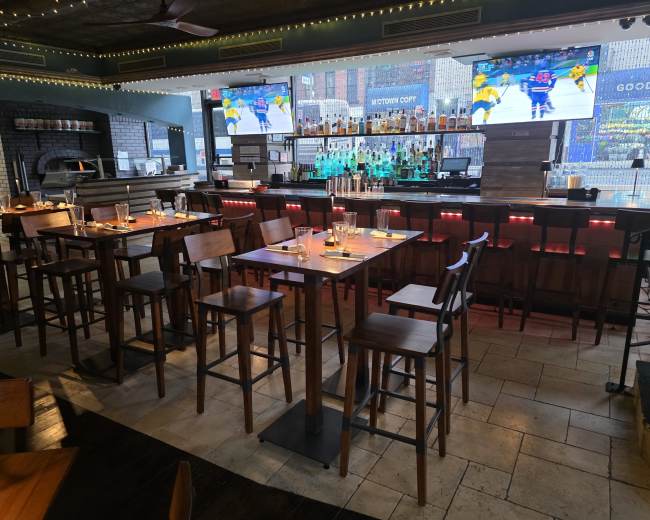 The image shows a modern bar area with high wooden stools, a well-stocked bar featuring multiple bottles, and large screens displaying a sports game in a spacious establishment.