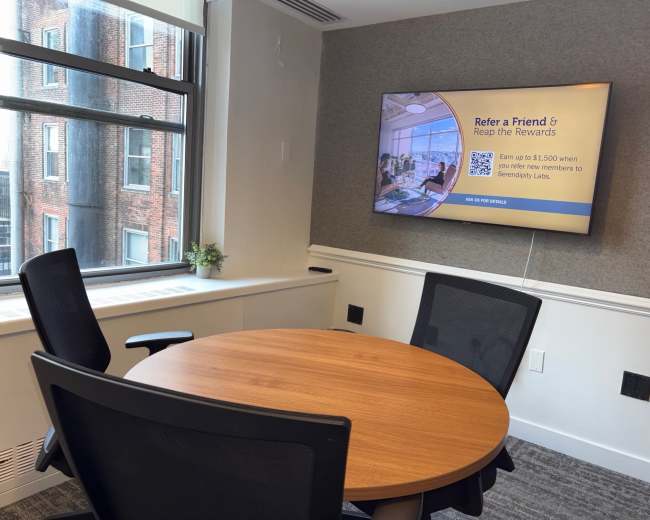 A conference room features a round wooden table, two black chairs, and a wall-mounted screen displaying a referral promotion.